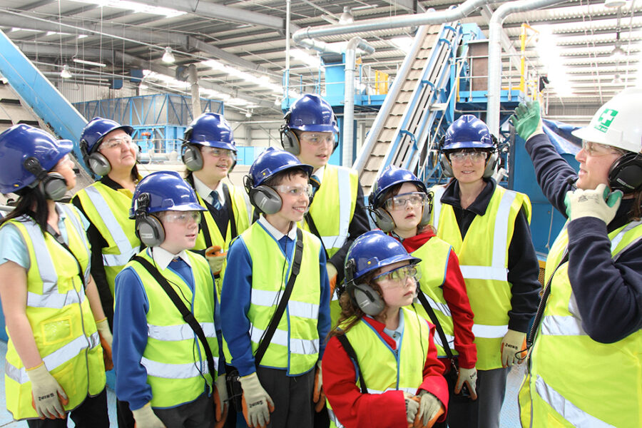 A school group being given a tour of a materials recovery facility. Everyone is wearing high-vis jackets, hard hats and ear defenders.