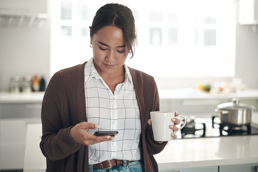 Shot of a young woman using a smart phone while drinking coffee in the kitchen at home.