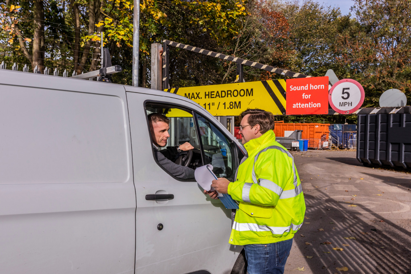 man in high vis jacket with clipboard speaks to man in van at the entrance of a recycling centre.