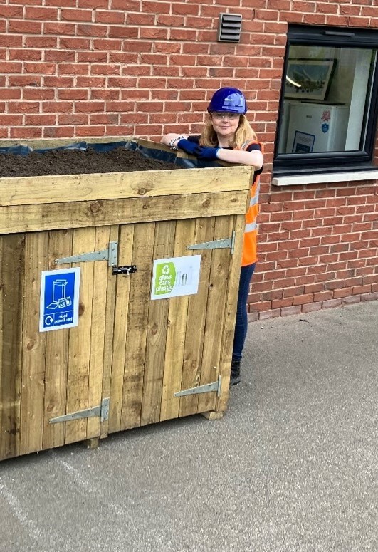 A woman in high vis and a hardhat leans on a wooden bin store which has a planter filled with soil on as a roof