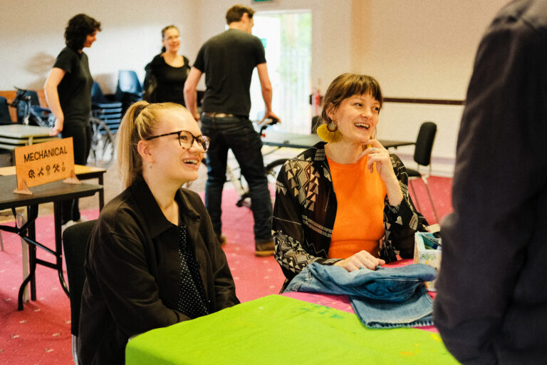 2 seated women smile as they work to repair a pair of jeans