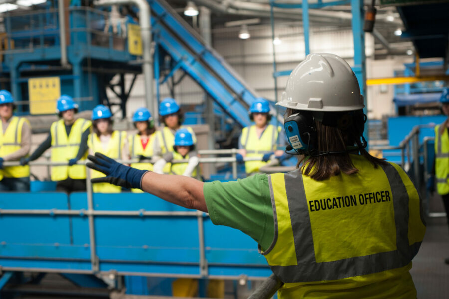photo of an education officer in high-vis and hard-hat giving a tour of the Materials recovery facility to a group of visitors